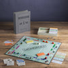 The Monopoly Vintage Bookshelf Edition is set up on a wooden table with premium cards, play money, dice, tokens, houses, and hotels. Its box, designed to look like a hardcover book, stands upright behind the game.