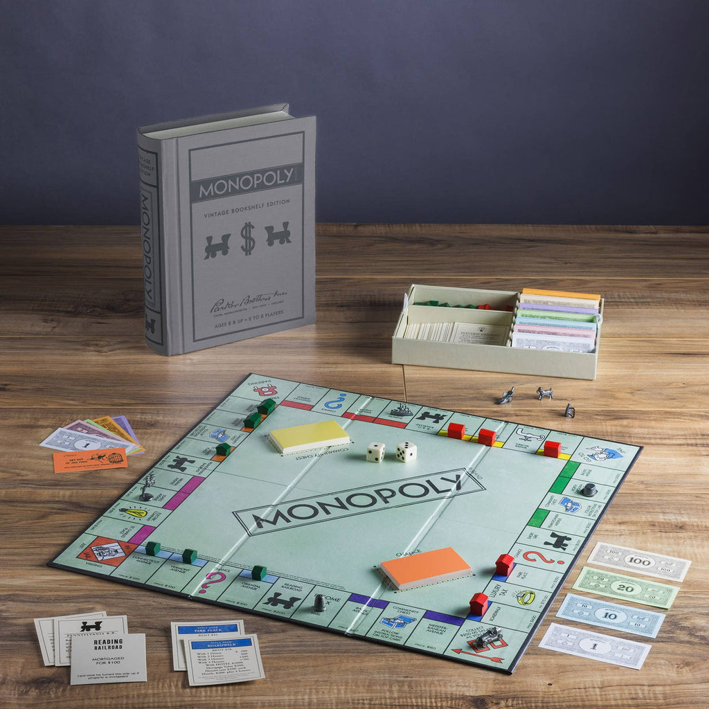 The Monopoly Vintage Bookshelf Edition is set up on a wooden table with premium cards, play money, dice, tokens, houses, and hotels. Its box, designed to look like a hardcover book, stands upright behind the game.
