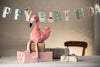 The Steiff Flamingo Stuffed Animal sits atop wrapped gifts on a table with a "Happy Birthday" banner in the background, adding charm to the festive scene and a wooden chair partially visible.