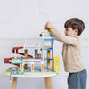 A young boy plays imaginatively with the Le Grand Wooden Garage, placing a car on the top level. This educational toy features ramps, signs, and multiple levels, set on a small table against a white wall.