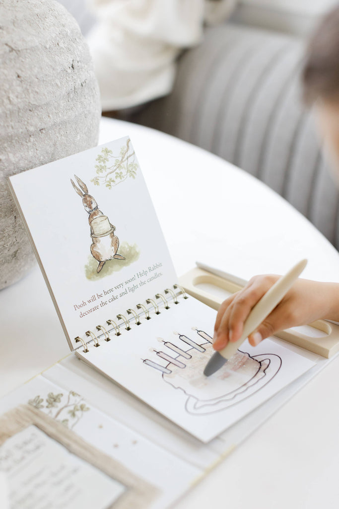 A child uses a water pen to reveal an illustration of a birthday cake on a Winnie-the-Pooh: Pooh’s Birthday Surprise (Water Reveal) spiral-bound book featuring a drawing of Rabbit.