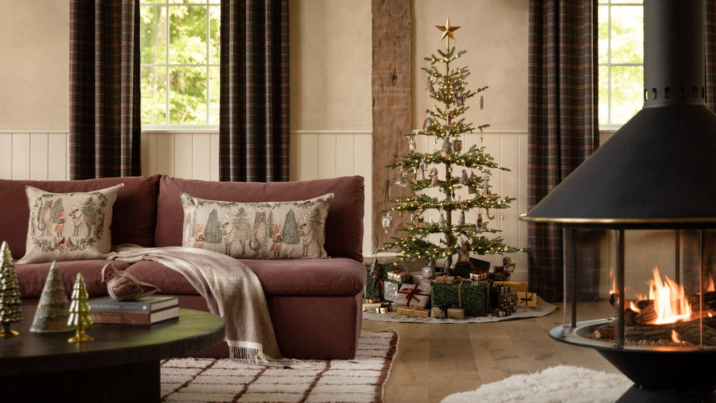 Coral & Tusk Christmas Cheer Wreath Pillow, a rectangular embroidered linen pillow featuring woodland animals and Christmas trees, displayed on a sofa next to a decorated Christmas tree.