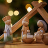 The Wooden Nativity Scene is handcrafted in Germany, showcasing the Holy Family and wise men beneath a simple wooden stable, softly illuminated by blurred festive lights in the background.