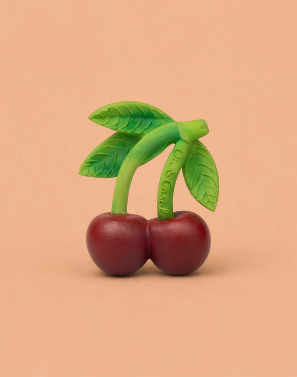 Mery the Cherry Teether is a realistic, rubbery teething toy shaped like two red cherries with green stems and leaves, made from natural rubber for sensory play, shown against a plain beige background.