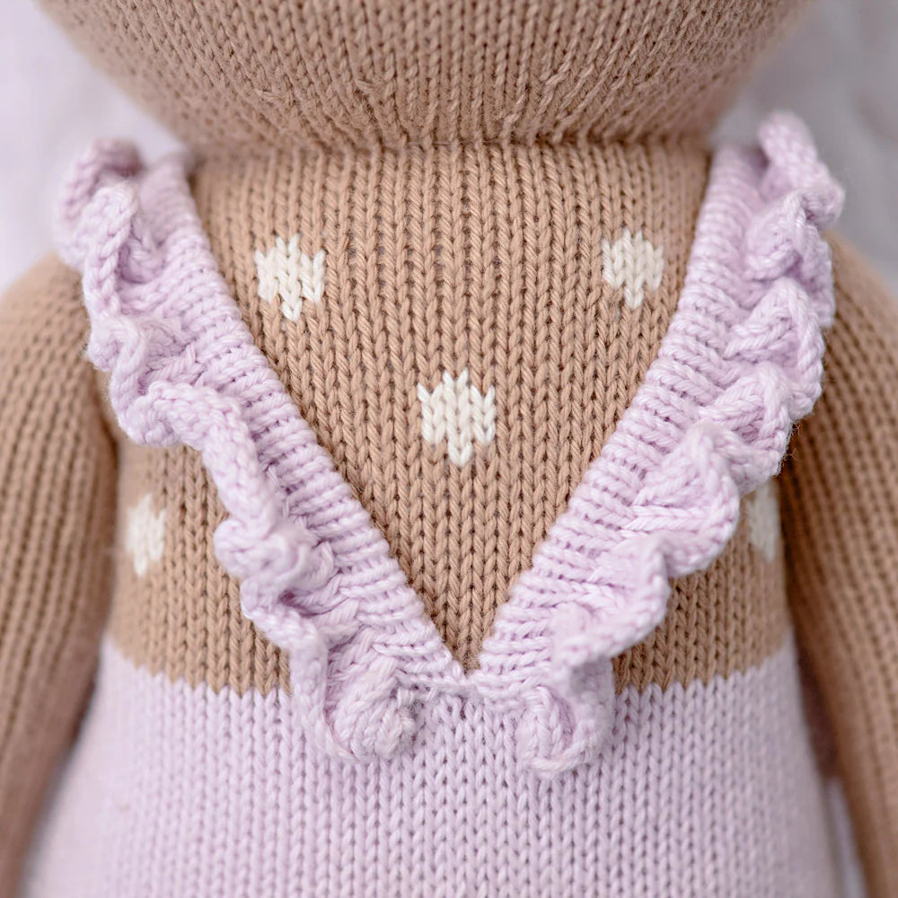 Close-up of Cuddle + Kind Violet the Fawn, a hand-knit doll in a light purple dress with ruffled straps and a V-shaped back. The light brown body features white heart-shaped patterns, crafted from premium cotton yarn.