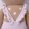 Close-up of Cuddle + Kind Violet the Fawn, a hand-knit doll in a light purple dress with ruffled straps and a V-shaped back. The light brown body features white heart-shaped patterns, crafted from premium cotton yarn.