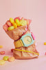 A brown paper bag filled with colorful round cereal pieces is wrapped with a Mini Kyomo Watch - Bubble Gum and secured with a pink rubber band, set against a pink background.