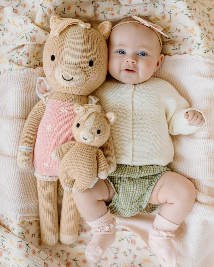 A baby lies on a floral blanket, dressed in a cream sweater, green checked shorts, pink socks, and a bow headband, holding two Cuddle + Kind Sophia the Pony dolls—one large and one small.