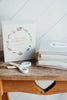 A box labeled "Our Little Library Vol. 3 (5 Board Books)" sits on a wooden bench beside titles "TOOLS," "SPORTS," "SPACE," and "DINOSAURS"—ideal for early language learning. A pair of white scissors rests nearby.