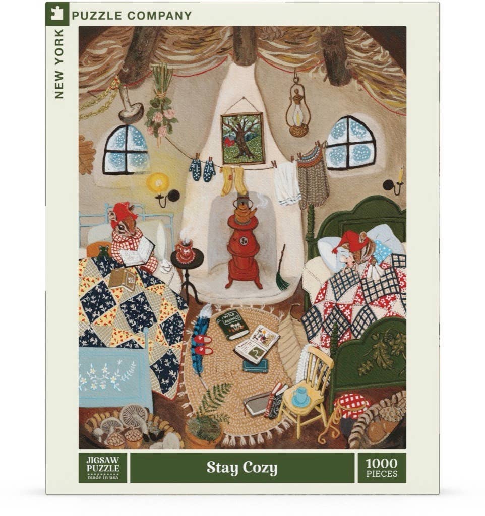 Stay Cozy - 1000 Piece Jigsaw Puzzle features two foxes relaxing in a rustic room with quilts, books, and a wood stove. A cozy puzzle and perfect holiday gift!.