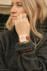A woman with blonde hair wears a black sweater, several rings, and the Mini Kyomo Watch - Cherry Bomb with a brown leather strap, looking to the side and resting her hand near her face.