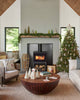 Cozy living room with a decorated Christmas tree, wrapped gifts, a round wooden coffee table, and a wood-burning stove. The mantel above the festive fireplace is adorned with holiday decorations and the Coral & Tusk Puppy Stocking Ornament.