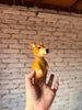 A hand holds up a small, handmade wool felt Shiba-Inu Dog Finger Puppet against a background of white brick wall and wooden ceiling, perfect for use as finger puppets.