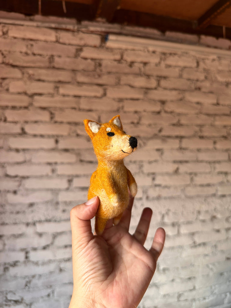 A hand holds up a small, handmade wool felt Shiba-Inu Dog Finger Puppet against a background of white brick wall and wooden ceiling, perfect for use as finger puppets.