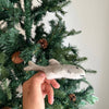 A hand holds the Finger Puppet: Shark, a small gray felt ornament, in front of a decorated Christmas tree with pinecones—adding imaginative underwater charm to your holiday decor.