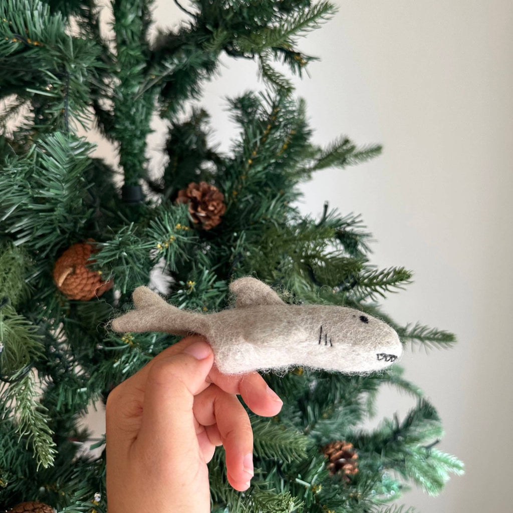 A hand holds the Finger Puppet: Shark, a small gray felt ornament, in front of a decorated Christmas tree with pinecones—adding imaginative underwater charm to your holiday decor.