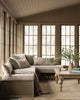 A cozy sunroom with large windows, a beige sectional bench featuring the Coral & Tusk Mushrooms and Ferns Lumbar Pillow among patterned and solid cushions, a wooden coffee table with books and greenery, plus firewood stacked beneath the bench.