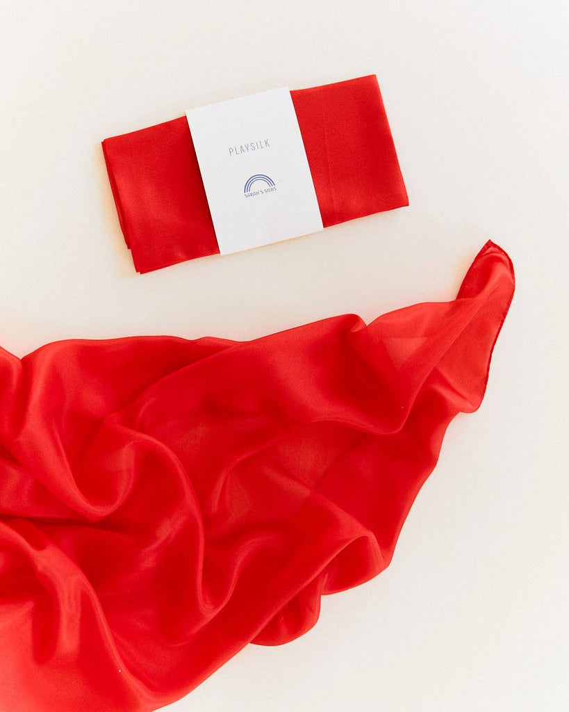 A folded Sarah's Silks Solid Color Playsilk - Red, made from pure silk and featuring a white label, rests above an unfurled red playsilk—ideal for open-ended play—against a light background.