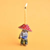 A whimsical Mushroom Gnome Cake Topper, hand painted, with a lit wick on top, set against a bright orange background.