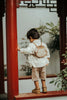 A young child with the Donsje Leather Kapi Backpack - Monkey stands facing away, touching a red pillar amid green plants and traditional architecture.