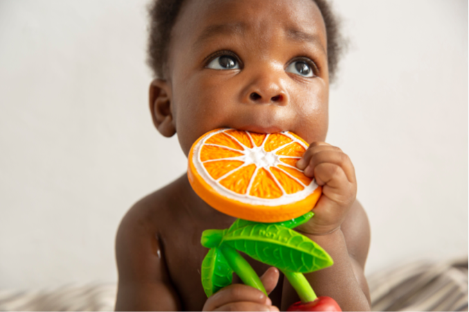 A baby looks upward while chewing on Clementino the Orange Teether, a natural rubber toy shaped like an orange slice with green leaves, against a neutral background.