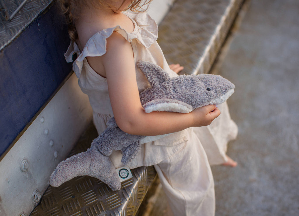 A young child in light clothing sits on metal stairs, holding a Senger Naturwelt Cuddly Animal - Shark plush. The child’s face is hidden, and soft natural light fills the scene.