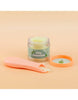 An open jar of "Tiny Tummies - Apple Jelly Food" by Tiny Harlow rests on a peach background, paired with a matching magic spoon. The jar’s label features an apple icon, enticing your child to relish the delicious flavor.