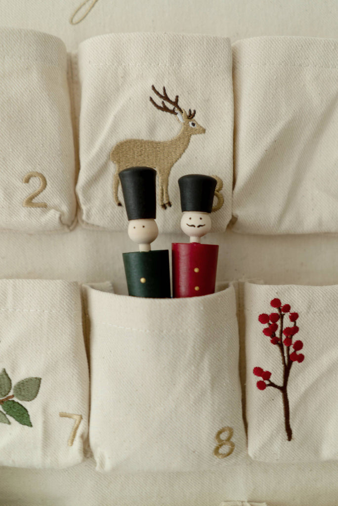 The textile advent calendar includes embroidered pockets adorned with numbers and festive motifs, such as a deer and red berries. Pocket number six contains two "Wooden Soldiers" peg dolls, one dressed in green and the other in red, both wearing small black hats.