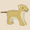 The Holztiger Lion Cub is a handcrafted wooden figure painted light brown with white accents, green eyes, and a cork-like tail. Made in Europe, it stands on all fours and ships in one week.