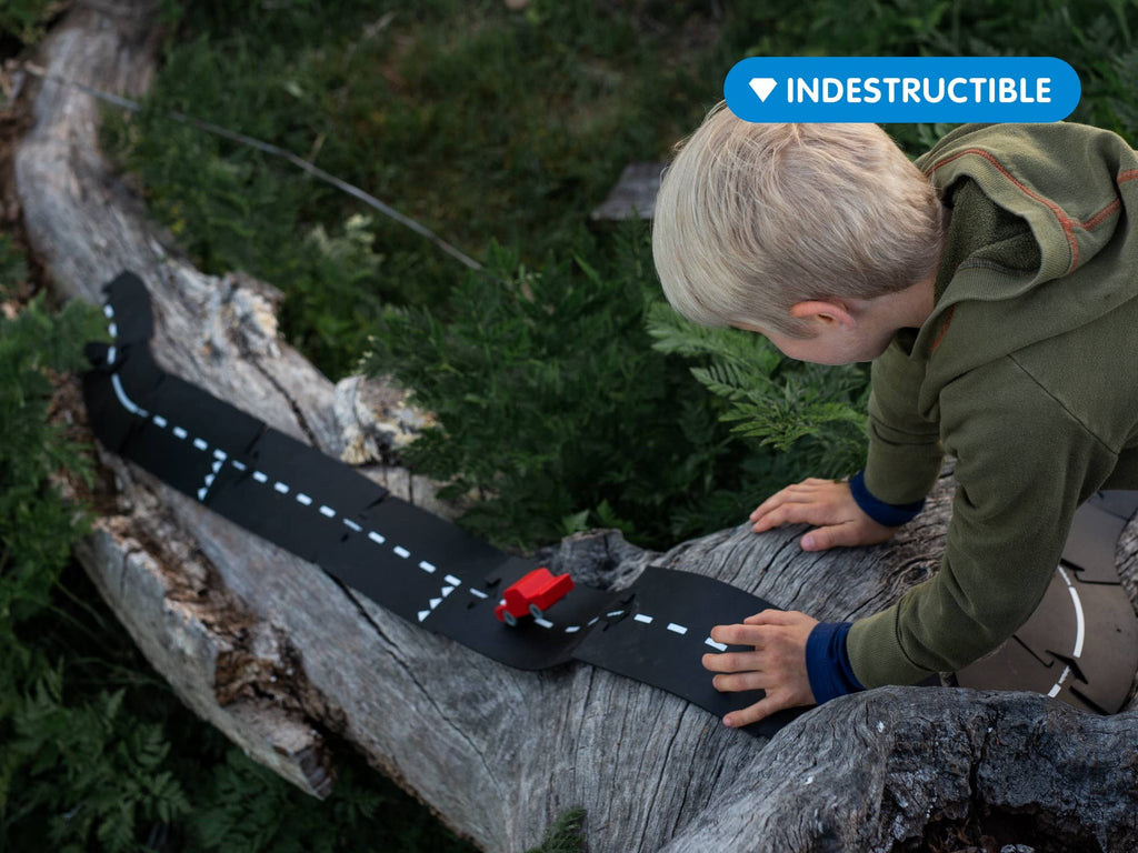 A young child plays outside, pushing a red toy car along the WaytoPlay: Medium Flexible Toy Road Set - Expressway, arranged on a fallen tree. A label in the corner states “Indestructible,” highlighting its durable, child-safe material.