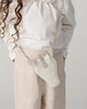 A child in a white blouse and beige pants holds the Donsje Leather Blinc Purse - Unicorn, a white leather purse with ears, horn, and an adjustable strap for comfort. Curly hair falls to one side.