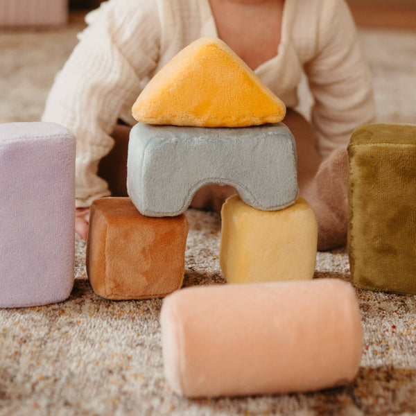 A baby plays on a carpet with Soft Toy Blocks—safe, non-toxic toddler toys in pastel colors like yellow triangle and light blue arch, inspired by Montessori blocks for fun and learning.