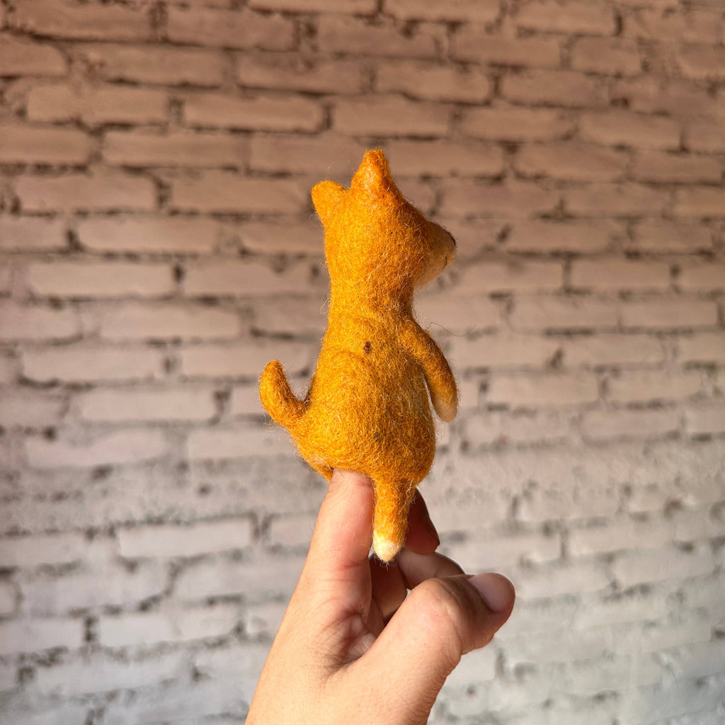 A hand holds a small, handmade orange felt Shiba-Inu Dog Finger Puppet with a tail, viewed from the back against a white brick wall—ideal for wool finger puppet enthusiasts.