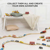 The Emergency Helicopter & Rescue Cars wooden toy set, featuring colorful rescue vehicles, is arranged in a wavy pattern on the floor. A bench with a pillow, cushion, and blanket sits behind. Text: "COLLECT THEM ALL AND CREATE YOUR OWN ADVENTURE.