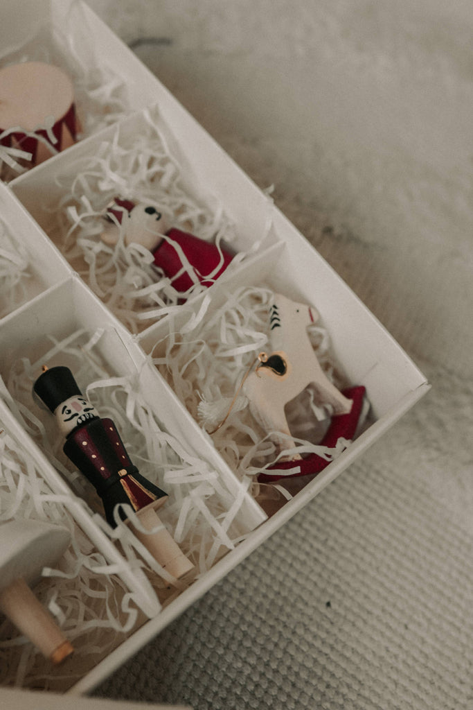 A close-up of a Sabo Concept Christmas Ornaments gift box shows compartments with white shredded paper, filled with hand-painted wooden decorations—a toy soldier, a white rocking horse, and a ballerina Christmas tree ornament.