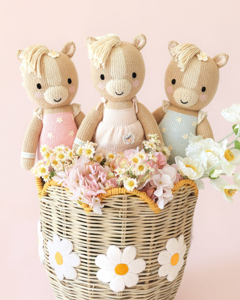 Three Cuddle + Kind Kelsie the Pony dolls in pastel dresses sit in a wicker basket with white daisies, surrounded by light pink and white flowers on a pale pink background. Each charming pony is lovingly handmade in Peru.