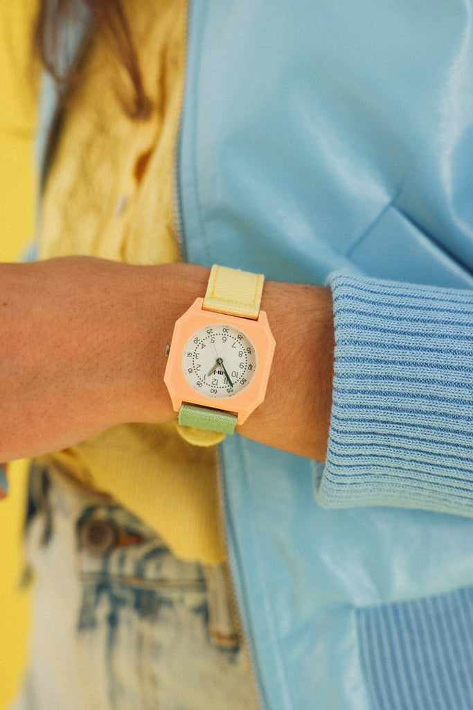 Someone wears the Mini Kyomo Watch - Watercolor in pastel yellow and green with a hexagonal peach face, paired with a light blue jacket and yellow top against a yellow background.