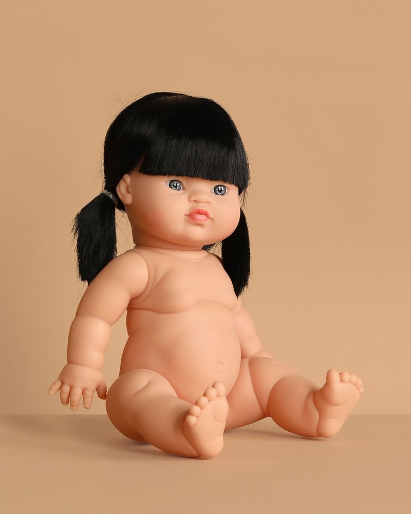 The Minikane Baby Doll (13") - Jade, featuring black pigtails and blue eyes, sits on a beige surface with a matching beige background. Clothing sold separately.