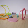 Grimm's Concentric Circles and Rings Building Set - Pastel features colorful wooden rings and disks scattered on a light wood surface, some upright, some flat or overlapping, ideal for building and balance games against a plain background.