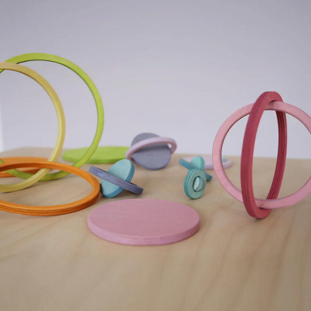 Grimm's Concentric Circles and Rings Building Set - Pastel features colorful wooden rings and disks scattered on a light wood surface, some upright, some flat or overlapping, ideal for building and balance games against a plain background.