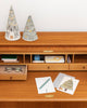 A wooden desk holds glasses, stationery, a teal pen, two cone-shaped keepsake decorations topped with Christmas tree and woodland creatures designs, and the Coral & Tusk Peek a Tree Card. Open cubbies below store notebooks and small items.