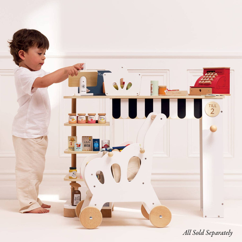 A small child plays pretend shopkeeper at a Le Toy Van wooden market stand with shelves, using the Cash Register & Money set, miniature groceries, and a white toy shopping cart in a bright, minimalist room.