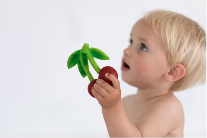 A blonde toddler looks up while holding the Mery the Cherry Teether, a safe baby toy, against a plain white background.