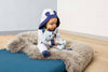 A baby in a hooded onesie with bear ears sits on a fluffy gray rug, cuddling the My First Steiff Blue Teddy Bear, 10 Inches, in a cozy room with wooden floors and white walls.