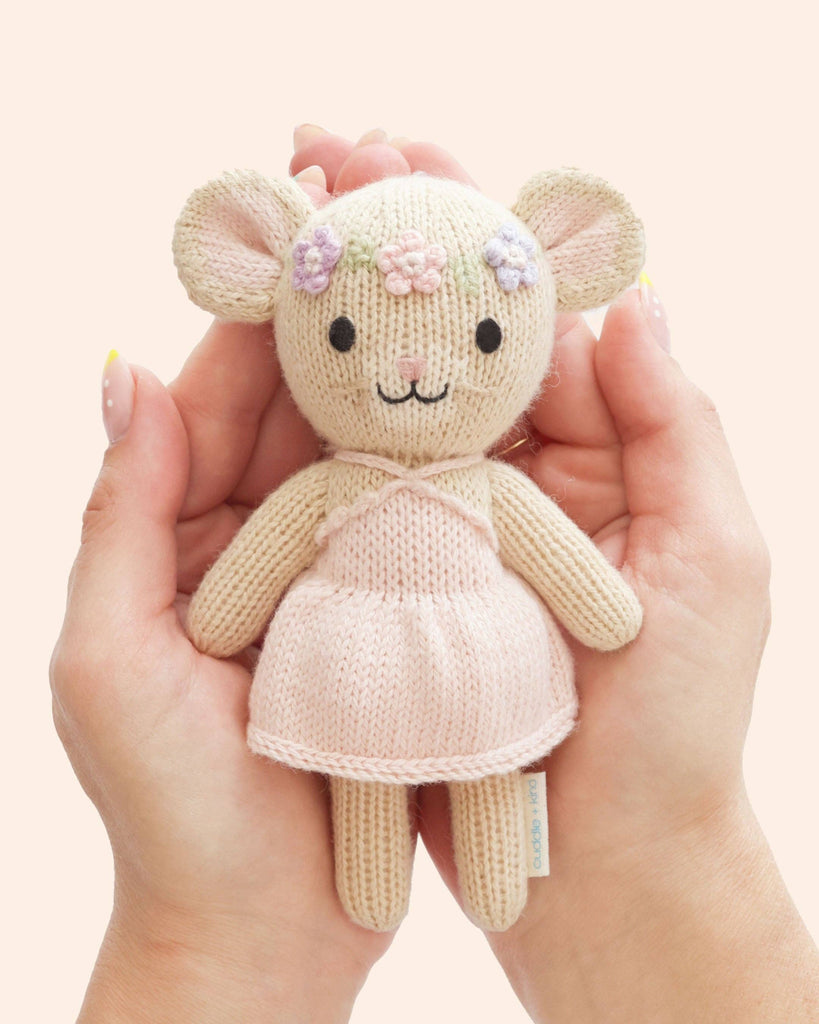 A pair of hands gently holds the Cuddle + Kind Tiny Emma the Mouse, a hypoallergenic, hand-knit stuffed mouse in a light pink dress with a flower crown, featuring a cute smile, round ears, and delicate embroidered details.