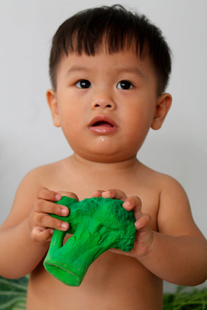A young child with short hair, shirtless, holds Brucy the Broccoli Teething Toy made from green natural rubber and looks up with a curious expression.