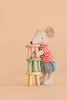 A stuffed mouse in a red striped shirt and blue skirt stands upright, stacking the Maileg Stool Set, Mouse – Light Rose/Light Mint, featuring small pastel-colored stools crafted from beech wood with a charming vintage paint finish, against a plain beige background.