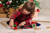 A young child in a red plaid dress plays with the BRIO World Christmas Steaming Train Set on the floor near a decorated Christmas tree and wrapped presents.
