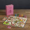 The Board Game: Candy Land Vintage Bookshelf Edition is displayed on a wooden table with colorful pieces, cards, and a bright winding board. The pink game box stands upright in the background, designed to look like a hardcover book.