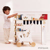 A young boy enjoys the Groceries Set & Scanner, featuring pretend shopping toys like a red cash register, scale, shelves of mini products, and a pushcart—all made from FSC-certified wood—in a bright white room.
